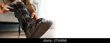 Ankle foot orthosis after injury. Man putting ankle orthosis on leg sitting on the sofa at home. Stock Photo