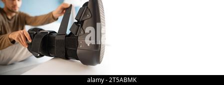 Man putting ankle sprain orthosis, black splint on leg sitting on the sofa on white background. Ankle foot orthosis. Banner. Stock Photo