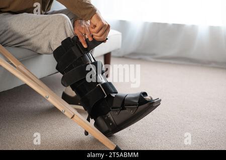 Young man with leg injury using crutch and orthosis sitting on sofa at home Stock Photo