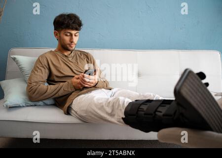 Man with leg injury lying on sofa with phone, ankle brace orthosis on male leg at home Stock Photo