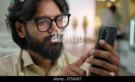 Indian male bearded man in glasses professional hold modern smartphone texting message in office indoors. Arabian businessman using mobile app Stock Photo