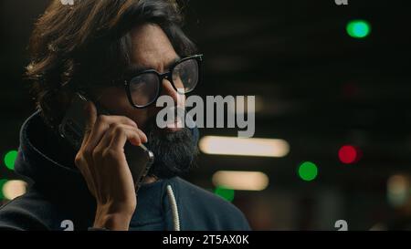 Smiling Arabian adult ethnic man guy in glasses casual businessman in eyeglasses making business call talking phone standing at dark parking lot Stock Photo