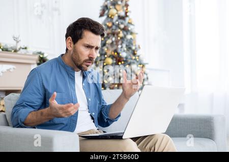 Frustrated sad and unhappy man sitting on sofa in living room near Christmas tree, businessman working remotely on New Year holidays, looking at laptop confused. Stock Photo