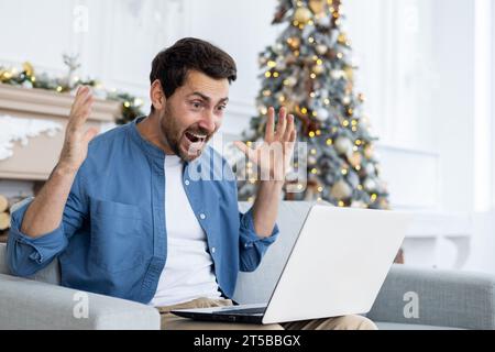 Man joyfully shouting at laptop screen got online win and victory, achievement results are good, celebrating triumph, sitting on sofa in living room, celebrating new year and christmas. Stock Photo