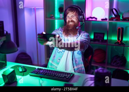 Young redhead man streamer playing video game using smartphone at gaming room Stock Photo