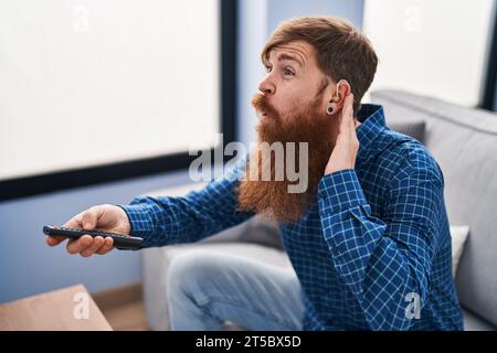 Young redhead man using deafness aid machine watching tv at home Stock Photo