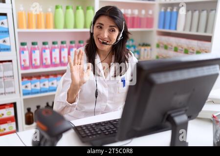 Middle age hispanic woman working at pharmacy drugstore using headset and computer looking positive and happy standing and smiling with a confident sm Stock Photo