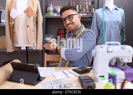 Hispanic man with beard dressmaker designer working at atelier inviting to enter smiling natural with open hand Stock Photo
