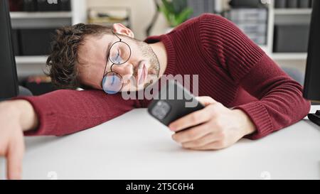 Young hispanic man business worker using smartphone tired sleeping on table at office Stock Photo