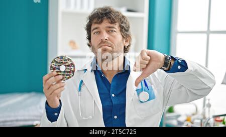 Young hispanic man nutritionist holding doughnut doing thumb down gesture at clinic Stock Photo