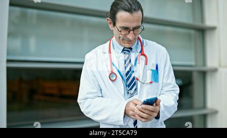 Middle age man doctor smiling confident using smartphone at hospital Stock Photo