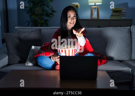Hispanic woman eating popcorn watching a movie on the sofa smiling cheerful offering palm hand giving assistance and acceptance. Stock Photo