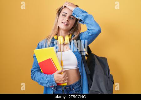 Young blonde woman wearing student backpack and holding books smiling confident touching hair with hand up gesture, posing attractive and fashionable Stock Photo