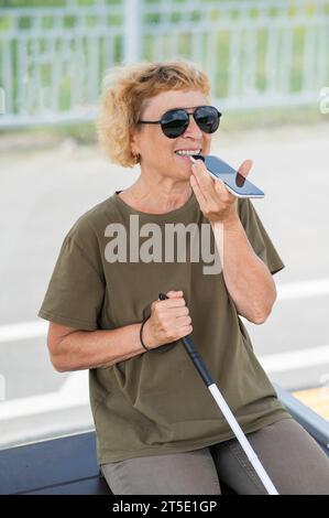 Elderly blind woman sitting at a bus stop and using a smartphone.  Stock Photo