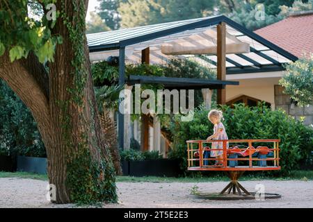 Little girl stands on a carousel in a green garden. Side view Stock Photo