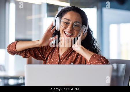 Young beautiful woman working in the office at the workplace, close-up hispanic woman in headphones singing along and dancing joyfully, business woman on break resting, programmer happy. Stock Photo