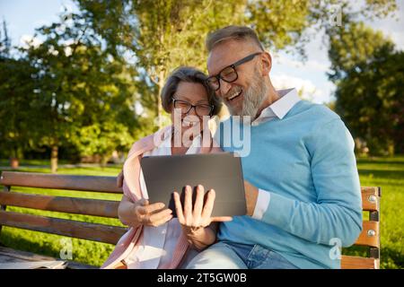Happy cheerful senior couple using digital tablet while rest on park bench Stock Photo