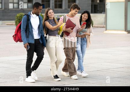 Young group of students walking and using phones in the street. Diverse students texting and laughing while moving in the campus. Stock Photo