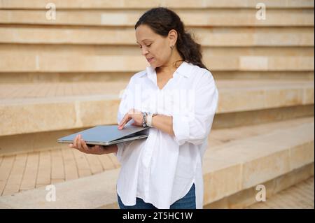 Confident authentic young multi ethnic woman in white casual shirt, opening laptop standing against beige marble steps background. People. Career. Onl Stock Photo