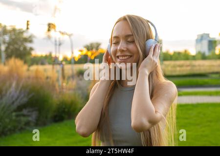 Cheerful young woman enjoys listening to music and singing while sitting in park. Happy lady in headphones rests spending time in nature Stock Photo