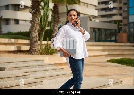 Portrait of charming young adult woman dressed in stylish white shirt and blue jeans, holding laptop and looking confidently into the distance while w Stock Photo