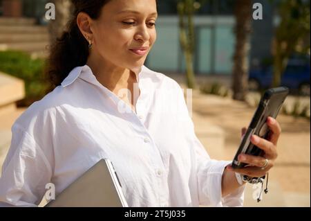 Portrait of beautiful young woman using smartphone, checking social media content or new mobile application, standing with laptop in her hands, dresse Stock Photo