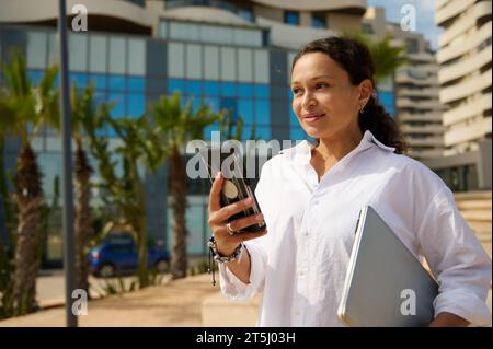 Portrait of young smiling dark-haired woman in casual white shirt walking city streets and using smartphone to interact with her business partners. Pe Stock Photo