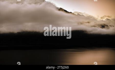 landscape photohraphy of the Lac du Mont Cenis in the French Alps during the golden hour, dense clouds covering the mountain tops, calm water, dark Stock Photo