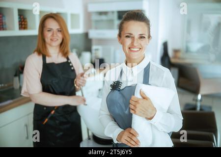 happy two 40 years old hair salon employees in aprons in modern hair studio near salon backwash unit. Stock Photo