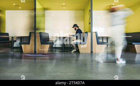 Software developer works on his laptop in a busy coworking office. Surrounded by business people, he focuses on coding and programming in his booth. V Stock Photo