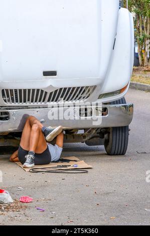 NEW ORLEANS, LA, USA - OCTOBER 29, 2023: Woman laying on her back under the front of a large truck replacing the fan belt. Stock Photo