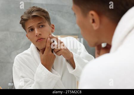 Upset young man looking at mirror and popping pimple on his face indoors. Acne problem Stock Photo