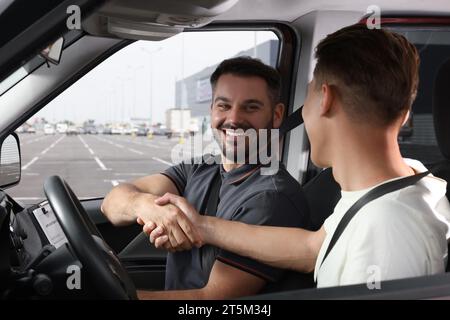 Driving school. Happy student shaking hands with driving instructor during lesson in car at parking lot Stock Photo