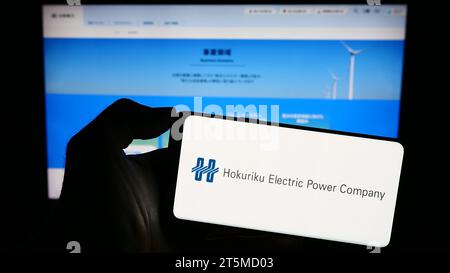 Person holding cellphone with logo of Japanese energy business Hokuriku Electric Power Company in front of webpage. Focus on phone display. Stock Photo