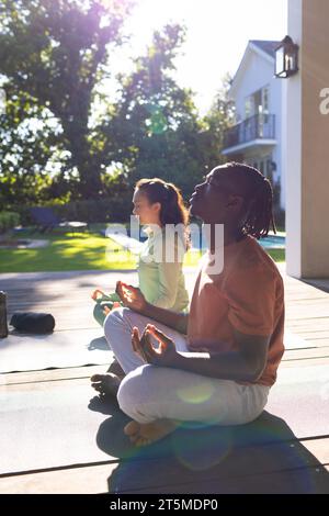 Happy diverse couple practicing yoga meditation sitting on deck in sunny garden, copy space Stock Photo