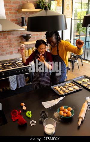 Happy diverse couple in aprons baking christmas cookies, dancing in kitchen Stock Photo