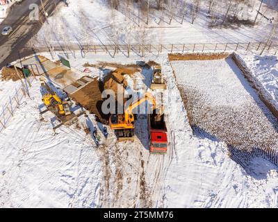 Aerial view of an excavator pouring sand into a truck. Top view of the construction site in winter. Shooting from a drone. Stock Photo