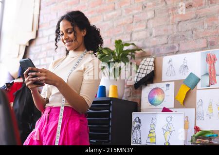 Happy biracial female fashion designer using smartphone and smiling in sunny studio Stock Photo