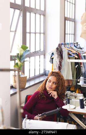Pensive biracial female fashion designer using tablet and looking out window in sunny studio Stock Photo