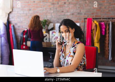 Focused biracial female fashion designer using smartphone and laptop in sunny studio Stock Photo