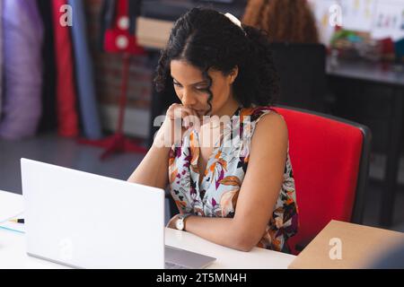 Focused biracial female fashion designer using laptop in sunny studio Stock Photo