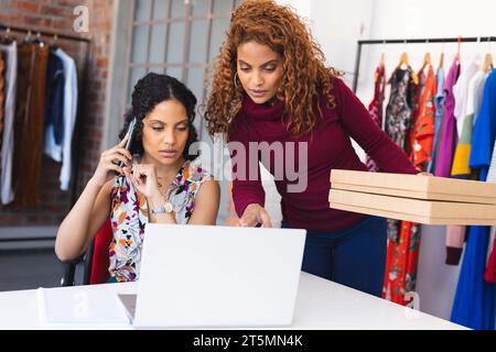 Two biracial female fashion holding packing parcels, using smartphone and laptop in sunny studio Stock Photo