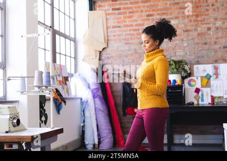 Focused biracial female fashion designer working with tablet in sunny studio Stock Photo