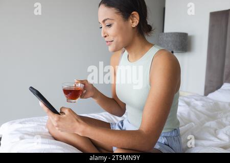 Happy biracial woman using smartphone and drinking tea in bedroom Stock Photo