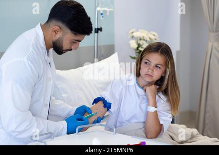 Focused diverse male doctor taking blood sample with syringe from girl patient in hospital bed Stock Photo