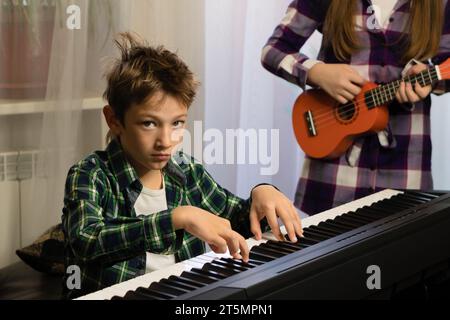 A boy intently plays piano while his sister practices the ukulele, creating a harmonious duet at home Stock Photo