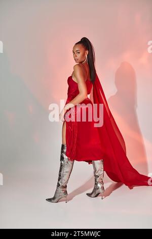 charming african american woman in red dress with shawl posing in silver boots on grey with lighting Stock Photo