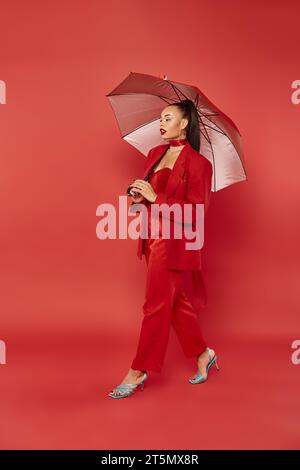 pretty african american woman in blazer and pants walking under umbrella on red backdrop Stock Photo