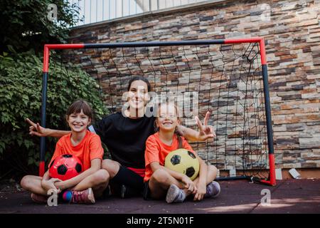 Mom playing football with her daughters, dressed in football jerseys. The family as one soccer team. Family sports activities outside in the backyard Stock Photo