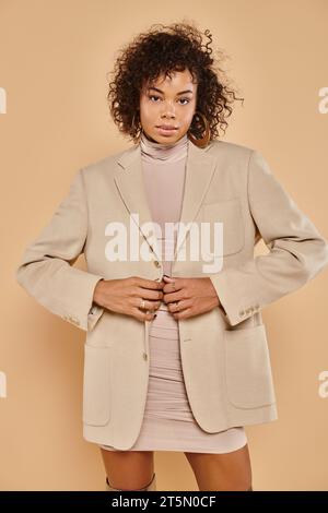 autumn fashion, stylish african american woman posing in tight dress and blazer on beige backdrop Stock Photo